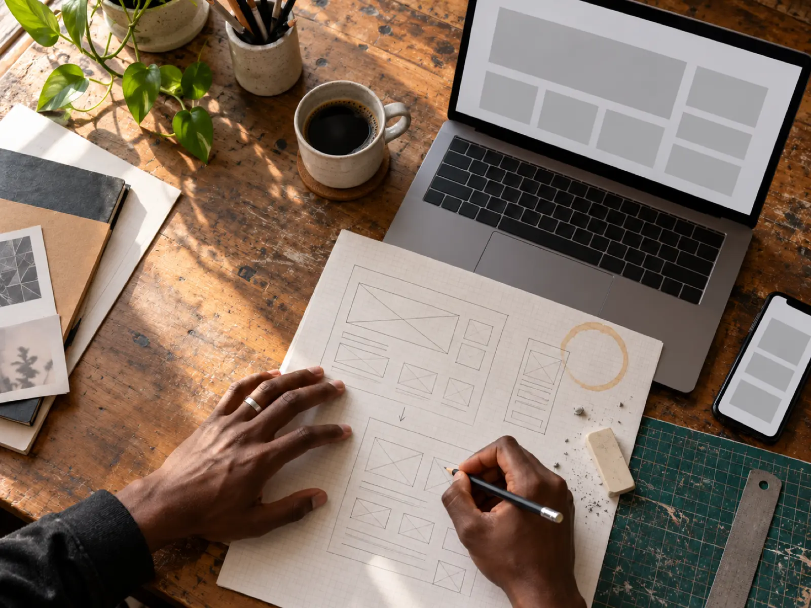Designer sketching homepage wireframes on paper beside a laptop showing a website layout